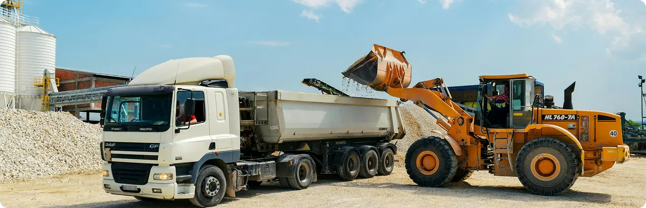 png 1 Camião basculante branco a ser carregado por máquina escavadora amarela num estaleiro de construção.